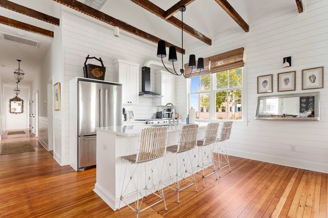 Bright coastal farmhouse kitchen with white shiplap walls, exposed dark wood ceiling beams, hardwood floors, stainless steel refrigerator, white island with four woven bar stools, black pendant chandelier, and a large window with tree views.