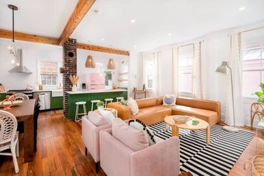 Bright open-concept living room and kitchen with tan leather sectional, pink retro fridge, green island, exposed wood beams, striped rug and hardwood floors.