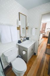 Chic small bathroom with marble vanity, black faucet, brass sconces, geometric wallpaper, white towels, toilet, and warm hardwood floor leading to an exposed-brick room.