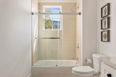 Bright modern bathroom with white bathtub and sliding glass shower doors, beige tile surround, window letting in natural light, white toilet and framed coastal wall art.