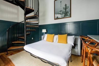 Cozy loft bedroom with a black metal spiral staircase, dark teal wainscoting, white bed with two yellow accent pillows, wooden floors, rattan bar stools, and Eiffel Tower wall art.
