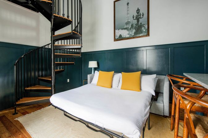 Cozy loft bedroom with a black metal spiral staircase, dark teal wainscoting, white bed with two yellow accent pillows, wooden floors, rattan bar stools, and Eiffel Tower wall art.