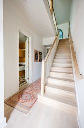 Bright modern home hallway with light oak staircase and white balustrade leading to an arched blue doorway, pale hardwood floors topped by a patterned southwestern runner, an open bedroom door showing bunk beds, and framed artwork on the wall.