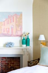 Coastal-inspired bedroom nook with white mantel over exposed brick, pastel house painting, turquoise vases with magnolia blooms, gold wall sconce and pale green pillow.