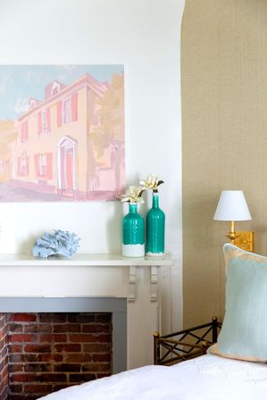 Coastal-inspired bedroom nook with white mantel over exposed brick, pastel house painting, turquoise vases with magnolia blooms, gold wall sconce and pale green pillow.