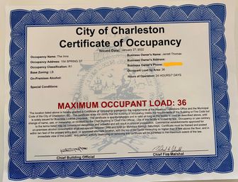 City of Charleston Certificate of Occupancy with blue scalloped border and faded seal watermark, bold red text reading "MAXIMUM OCCUPANT LOAD: 36", printed occupancy details and contact info (redacted), and official signatures at the bottom.