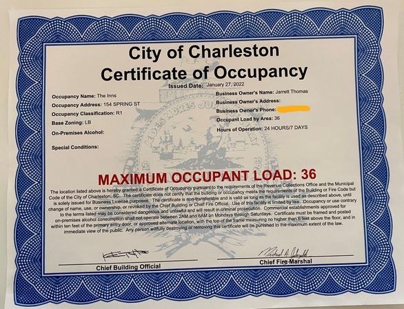 City of Charleston Certificate of Occupancy with blue scalloped border and faded seal watermark, bold red text reading "MAXIMUM OCCUPANT LOAD: 36", printed occupancy details and contact info (redacted), and official signatures at the bottom.