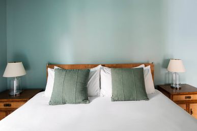 Cozy minimalist bedroom with white linens, two sage-green throw pillows, wooden headboard and matching nightstands with glass lamps against a seafoam green wall.