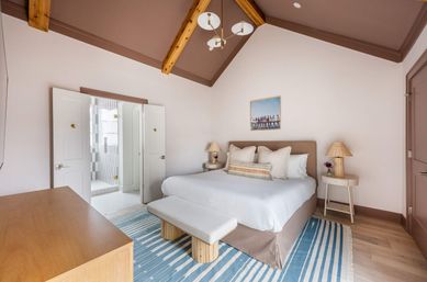Relaxing coastal-inspired master bedroom with vaulted wood-beamed ceiling, king bed with neutral linens, bench at foot, blue-striped rug, bedside lamps on round tables, and open double doors leading to a tiled en-suite bathroom.