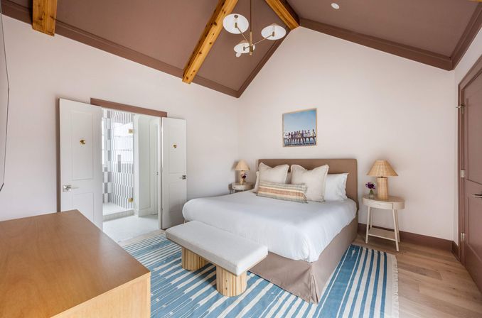 Relaxing coastal-inspired master bedroom with vaulted wood-beamed ceiling, king bed with neutral linens, bench at foot, blue-striped rug, bedside lamps on round tables, and open double doors leading to a tiled en-suite bathroom.