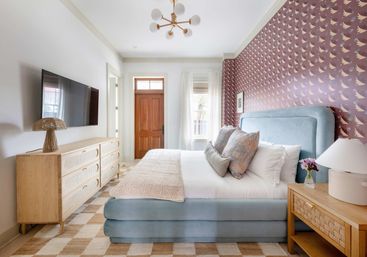 Airy modern bedroom with plush powder-blue upholstered bed, mauve bird-patterned wallpaper, light wood dresser and nightstand, checkered rug, wall-mounted TV, and natural light from a window and wooden door.