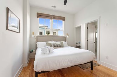 Bright, airy bedroom with king bed and beige headboard, white linens and folded towels, hardwood floors, twin windows with woven shades, wall sconces and door to en-suite bathroom.