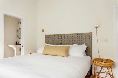 Bright minimalist bedroom with woven gray headboard, crisp white bedding, mustard lumbar pillow, rattan round side table, brass wall sconces, and an open doorway revealing a pedestal sink and round mirror