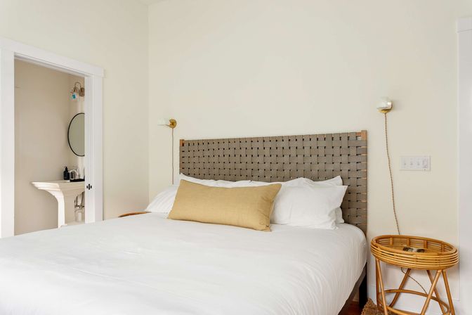 Bright minimalist bedroom with woven gray headboard, crisp white bedding, mustard lumbar pillow, rattan round side table, brass wall sconces, and an open doorway revealing a pedestal sink and round mirror