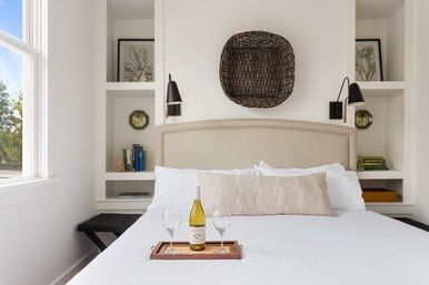 Sunlit minimalist bedroom with white bedding, beige upholstered headboard, woven basket wall decor, black wall sconces and built-in shelves; wooden tray with a bottle of white wine and two glasses on the bed.