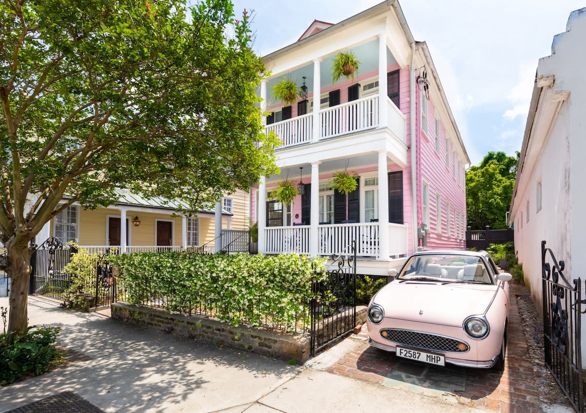 Charming two-story pink historic home with white double porches, hanging ferns, black shutters, iron fence with ivy, and a pastel pink vintage convertible parked in the driveway on a tree-lined southern neighborhood street.