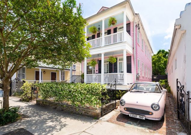 Charming two-story pink historic home with white double porches, hanging ferns, black shutters, iron fence with ivy, and a pastel pink vintage convertible parked in the driveway on a tree-lined southern neighborhood street.