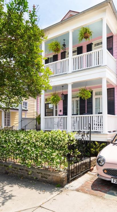 Charming two-story pink historic home with white double porches, hanging ferns, black shutters, iron fence with ivy, and a pastel pink vintage convertible parked in the driveway on a tree-lined southern neighborhood street.