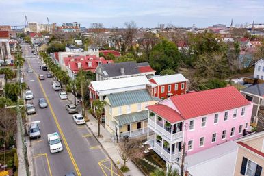 Aerial view of a historic coastal downtown street with colorful pastel homes and red metal roofs, tree-lined sidewalks, parked cars, and a distant suspension bridge.