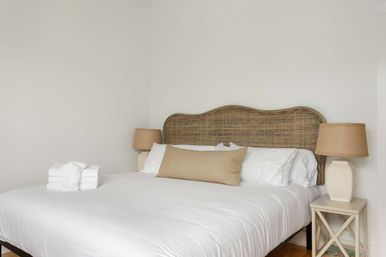 Neutral minimalist bedroom with woven rattan headboard, crisp white bedding, beige lumbar pillow, matching bedside lamps and neatly folded white towels on the bed