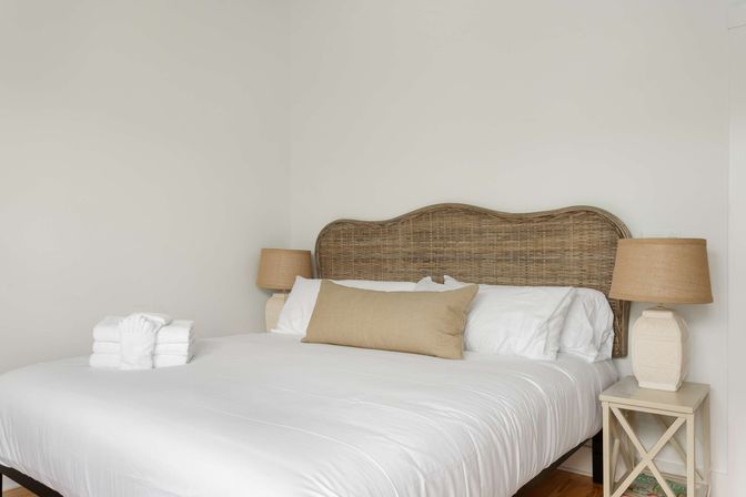 Neutral minimalist bedroom with woven rattan headboard, crisp white bedding, beige lumbar pillow, matching bedside lamps and neatly folded white towels on the bed