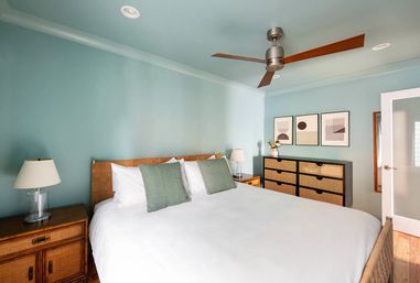 Coastal-inspired bedroom with teal walls and ceiling, king bed dressed in white linens and green accent pillows, rattan headboard and nightstands, wooden-blade ceiling fan, woven-rattan dresser, abstract wall art and a small vase of flowers.