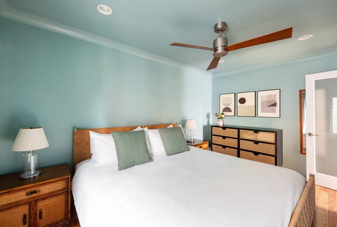 Coastal-inspired bedroom with teal walls and ceiling, king bed dressed in white linens and green accent pillows, rattan headboard and nightstands, wooden-blade ceiling fan, woven-rattan dresser, abstract wall art and a small vase of flowers.