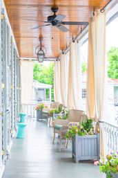 Bright covered porch with wooden ceiling, wicker chairs with cushions, flowing cream curtains, ceiling fans and lanterns, and colorful flower-filled planters