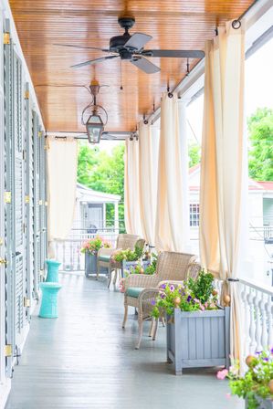 Bright covered porch with wooden ceiling, wicker chairs with cushions, flowing cream curtains, ceiling fans and lanterns, and colorful flower-filled planters