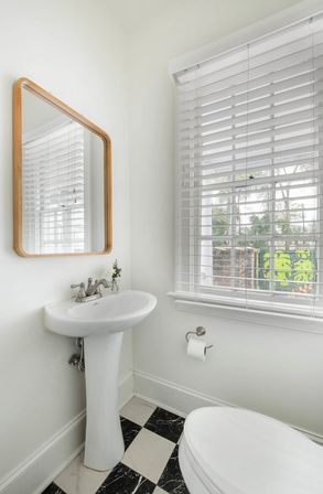 Bright white powder room with pedestal sink, wood-framed mirror, white horizontal blinds over a window with greenery, chrome faucet and toilet paper holder, small vase of flowers, and black-and-white checkered marble floor