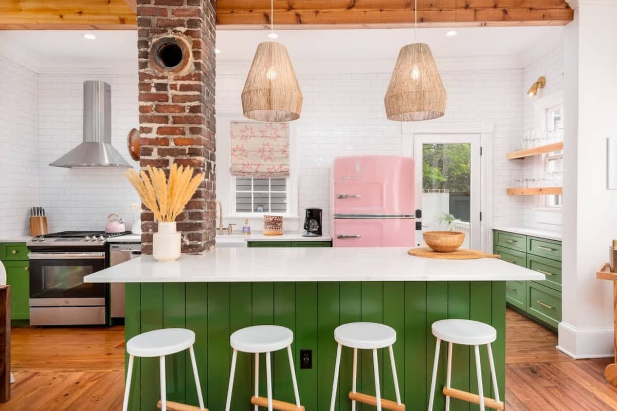 Bright retro farmhouse kitchen with green paneled island and white countertop, four white stools, exposed brick column, pink vintage refrigerator, stainless steel range, wicker pendant lights, white subway tile walls and hardwood floors.