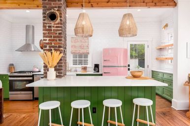 Bright retro farmhouse kitchen with green paneled island and white countertop, four white stools, exposed brick column, pink vintage refrigerator, stainless steel range, wicker pendant lights, white subway tile walls and hardwood floors.