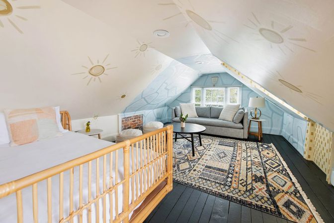 Cozy converted attic bedroom and lounge with sloped ceiling painted with sun motifs, rattan bed, gray sofa, patterned boho rug, round black coffee table and shuttered window