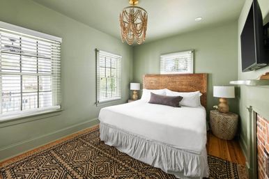 Cozy sunlit bedroom with sage-green walls, carved wooden headboard and king bed dressed in crisp white bedding and a dark lumbar pillow, woven geometric rug on hardwood floor, two windows with white blinds, beaded chandelier, bedside lamps, wall-mounted TV and brick fireplace.