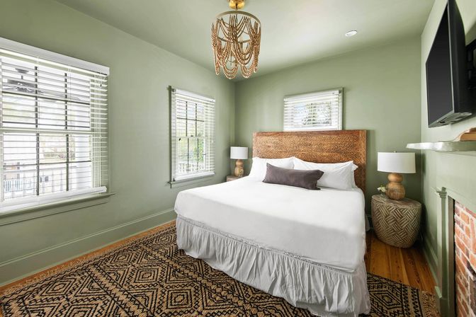 Cozy sunlit bedroom with sage-green walls, carved wooden headboard and king bed dressed in crisp white bedding and a dark lumbar pillow, woven geometric rug on hardwood floor, two windows with white blinds, beaded chandelier, bedside lamps, wall-mounted TV and brick fireplace.