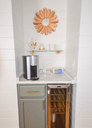 Cozy kitchen coffee station in a white shiplap niche with marble countertop, silver single-serve coffee maker, glass shelf holding mugs and gold accents, gray cabinet with brass hardware and a built-in wine fridge, topped by a decorative sunburst mirror.