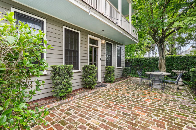 Cozy brick-paver patio beside a gray wood‑sided house with black‑trim windows, clipped shrubs, a round metal bistro table and chairs beneath a mature shade tree and privacy hedge.