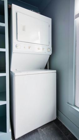 Neat white stacked washer-dryer in a compact laundry nook with pale blue walls, built-in shelving, tile floor and a nearby window.