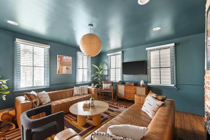 Cozy mid-century modern living room with teal walls and ceiling, tan leather sectional, round marble coffee table, rattan pendant light, wood floors, large windows with blinds, indoor plant and wall-mounted TV