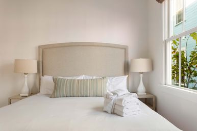 Sunlit minimalist bedroom with beige upholstered headboard, crisp white linens, striped lumbar pillow, neatly stacked white towels on the bed, matching ceramic bedside lamps and a window with green foliage outside.
