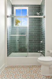 Bright modern bathroom with glass sliding shower over tub, teal subway tile surround, decorative floral-patterned floor tiles, white toilet and window showing blue sky.