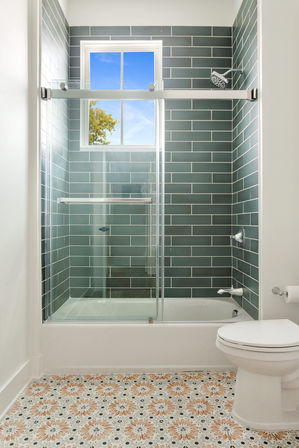 Bright modern bathroom with glass sliding shower over tub, teal subway tile surround, decorative floral-patterned floor tiles, white toilet and window showing blue sky.