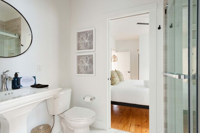 Sunlit modern white ensuite bathroom with pedestal sink, round mirror, glass shower door, framed coral prints, and an open doorway showing a white bed on warm hardwood floors