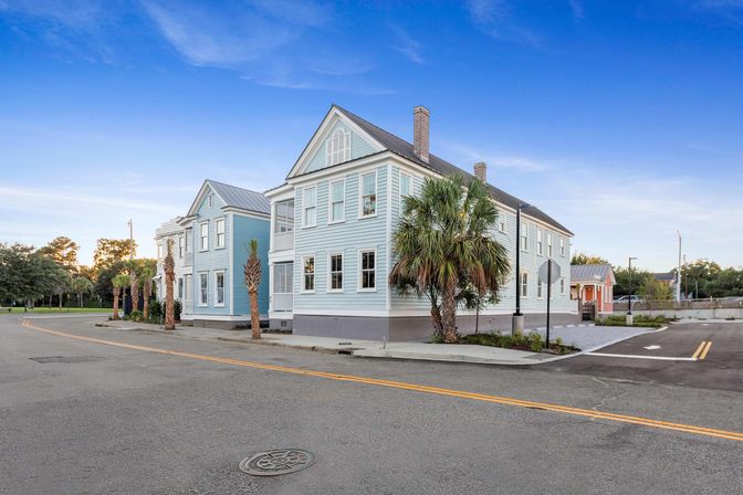 Light-blue historic-style homes on a sunny corner with palm trees, wide empty street and a clear blue sky — quaint coastal neighborhood scene.