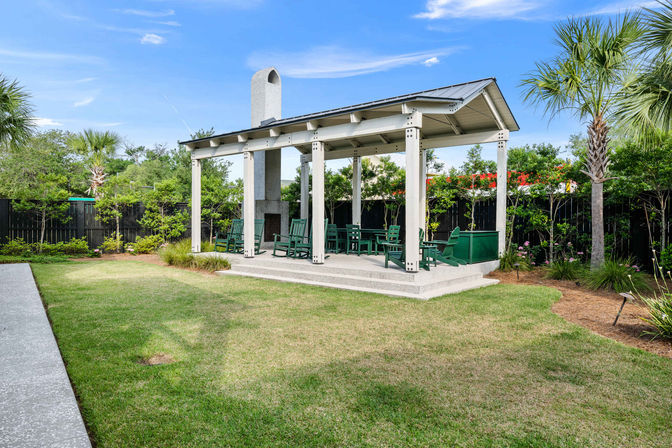 Covered backyard pavilion with tall outdoor fireplace, green wooden chairs on a concrete patio, manicured lawn and palm trees under a blue sky