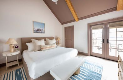 Bright coastal-style bedroom with vaulted ceiling and exposed wooden beams, plush king bed with neutral pillows, striped blue rug, bedside lamps on round tables, and French doors opening to a sunny balcony with water view.