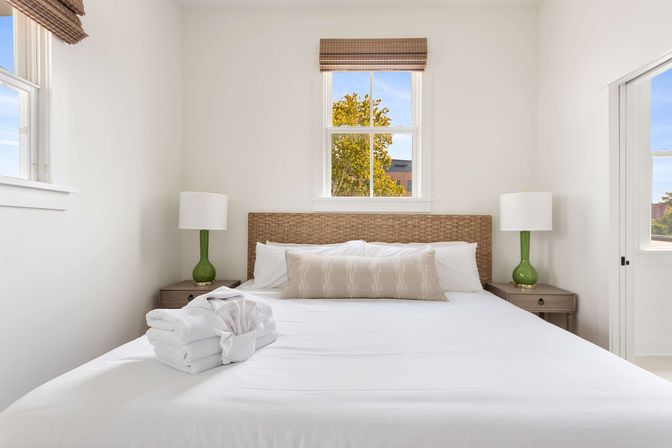 Sunlit minimalist bedroom with wicker headboard, crisp white linens and folded towels, green bedside lamps on wood nightstands, and a window showing a tree outside.