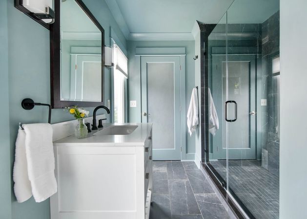 Spa-like modern bathroom with teal walls, white vanity and black matte fixtures, glass walk-in shower, gray stone floor and a small vase of yellow flowers.