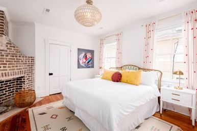 Bright coastal cottage bedroom with king bed in crisp white linens, yellow and red accent pillows, exposed brick fireplace, woven rattan basket, patterned area rug, brass bedside lamps, and two sunny windows with dotted curtains.