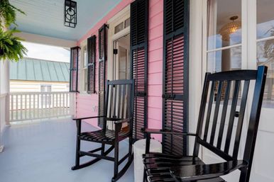 Cheerful pink front porch with pale-blue ceiling, black shutters and two black rocking chairs beside a hanging fern.